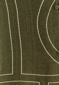 Tessuto verde con motivi di cucitura bianchi che formano linee e curve. La texture appare liscia con una leggera lucentezza. Dettaglio con bottone visibile.