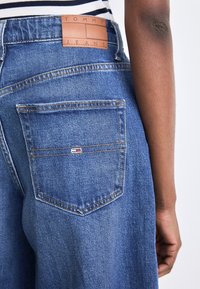 Blå denimjeans med en läderlogotyp på midjebandet och en bakficka med en liten logotags.