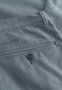 Tessuto grigio intrecciato con un motivo testurizzato, dotato di una tasca frontale e un dettaglio in bottone nero. Il design presenta rifiniture con cuciture ordinate.