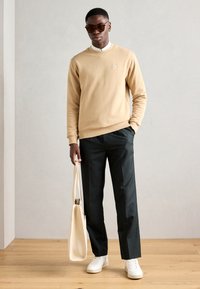 Sudadera beige con puños de canalé, pantalones negros, zapatillas blancas y una bolsa tote beige. La modelo lleva gafas de sol oscuras. Texturas suaves.