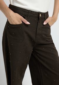 Pantaloni in denim marrone con sottili dettagli a borchie, design classico a gamba dritta e tasche frontali.