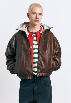 Mlajša oseba s kratkimi blond lasmi, oblečena v rjavo usnjeno jakno čez rdeč kardigan in črtasto majico, stoji na enobarvnem ozadju.