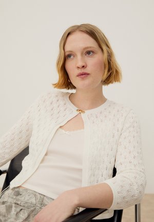 Femme aux cheveux blonds courts portant un cardigan blanc à motifs par-dessus un haut blanc, assise devant un fond clair uni.