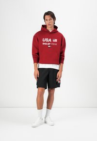 Rød hættetrøje med teksten "USA SAILGP TEAM" og flaggrafik; parret med sorte shorts og hvide sneakers. Simpelt design, afslappet pasform.