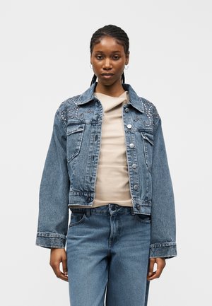Femme portant un haut beige clair, une veste en denim bleu avec des perles décoratives sur les épaules, et un jean bleu, debout devant un fond uni.