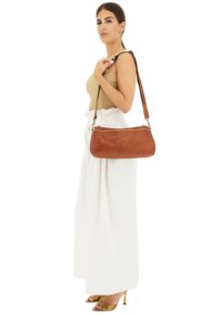 Borsa a tracolla in pelle marrone con forma arrotondata, dettaglio con borchie lungo la parte superiore e tracolla regolabile. Indossata sulla spalla.