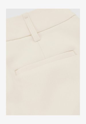 Tuvplāns uz bēšām auduma biksēm, redzama aizmugurējā kabata un jostas cilpa ar sīkām redzamo šuvju detaļām.