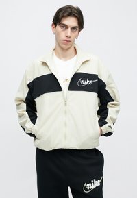 Αντιανεμικό μπουφάν Nike σε μπεζ και μαύρο, με πλήρες φερμουάρ, ελαστικά μανίκια και κεντημένο λογότυπο στο στήθος. Ομαλή υφή, άνετη εφαρμογή.