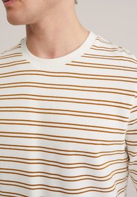 Person iført hvid T-shirt med rund hals og tynde vandrette brune striber, der viser nakke og nedre del af ansigtet i nærbillede.