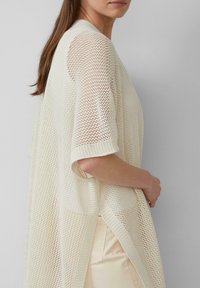 Cardigan de couleur crème, en tricot ouvert, avec des manches courtes, offrant une coupe décontractée et des fentes latérales. La texture en tricot est légère et respirante.