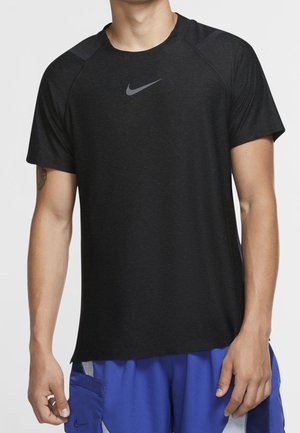 Mann trägt ein schwarzes Nike kurzärmeliges Sportshirt und blaue Shorts mit Seitentasche, steht vor einem schlichten Hintergrund.