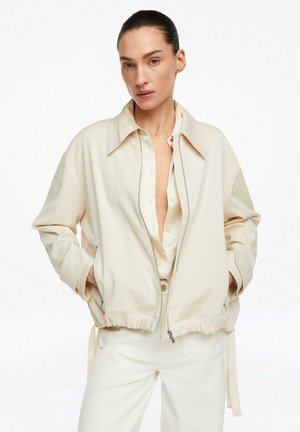 Kobieta ubrana w kremową kurtkę na częściowo rozpiętej koszuli i kremowe spodnie, stoi z rękami w kieszeniach kurtki na białym tle.