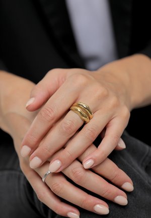 Guld bølget designring på en finger, ledsaget af en sølvring. Hænderne har lyserød neglelak, glat hud og let tekstur.