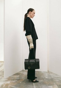 Geantă structurată neagră cu mâner de sus, material din piele netedă, asortată cu mănuși crem și haine negre minimaliste, pe un fundal deschis.