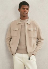 Bēšas krāsas jaka ar rāvējslēdzēju, ribveida akcentiem un divām priekšējām kabatām, nēsāta pāri gaiši bēšai kreklam, kopā ar krēmkrāsas biksēm. Gluda tekstūra.