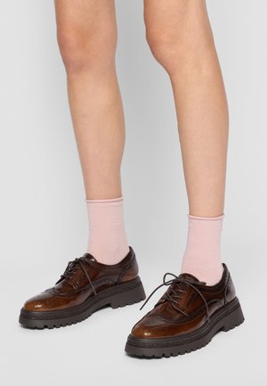 Des jambes portant des chaussettes rose clair et des chaussures brogue en cuir marron brillant avec des semelles épaisses noires, debout sur un sol gris clair.