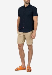 Dunkelblaues kurzärmliges Hemd mit Knopfleiste und einer Brusttasche sowie roten Knopfdetails, kombiniert mit hochgekrempelten khakifarbenen Shorts und orangenen Sandalen.