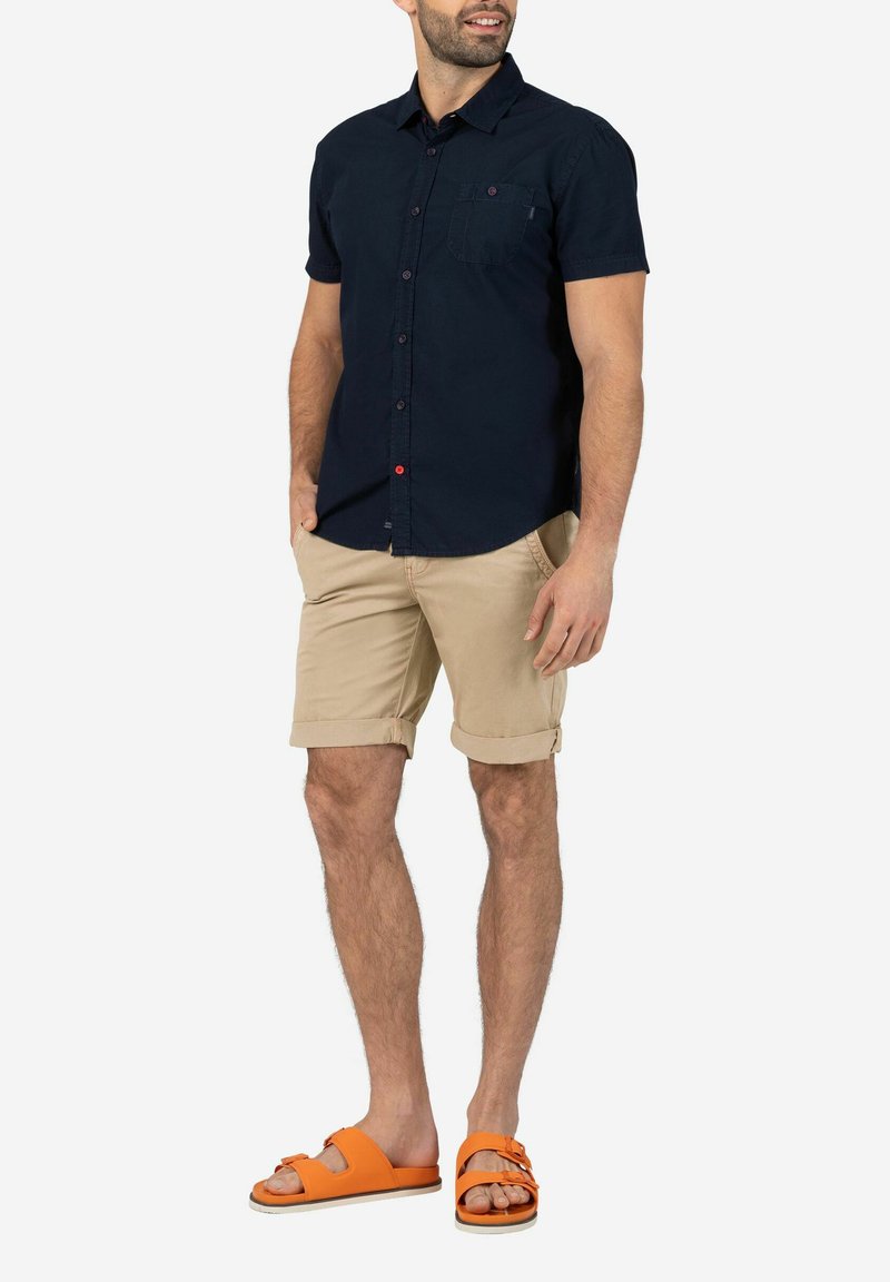 Dunkelblaues kurzärmliges Hemd mit Knopfleiste und einer Brusttasche sowie roten Knopfdetails, kombiniert mit hochgekrempelten khakifarbenen Shorts und orangenen Sandalen.