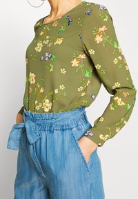 Oliwkowa bluzka w kwiatowe wzory w kolorze żółtym, niebieskim i białym. Długie rękawy, okrągły dekolt, połączona z dżinsowymi, wysokim stanem spodniami.