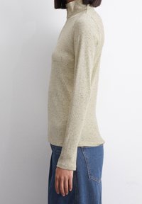 Kobieta ubrana w jasnobrązowy sweter z golfem w drobne kropeczki, z długimi rękawami i niebieską dżinsową spódnicą, stoi bokiem na jednolitym tle.