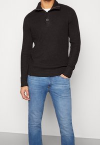 Man i svart ribbat pulloverwtröja med knappkrage och ljusblå jeans, stående mot en enkel bakgrund.