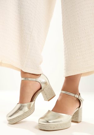 Scarpe con tacco a blocco dorato metallizzato e cinturini alla caviglia indossate con pantaloni ampi a gamba larga di colore bianco sporco e trama testurizzata.