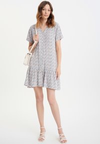 Vestido de manga corta con estampado floral en tejido ligero, con escote en V y dobladillo con volantes. La modelo sostiene un pequeño bolso blanco.