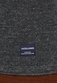 Dunkelgrauer Strickstoff mit einem kleinen marineblauen Etikett, auf dem "JACK & JONES Authentic Jeanswear Since 1990" steht, über braunem Stoff.