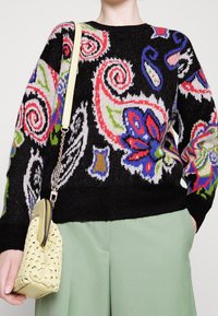 Pull noir avec des motifs cachemire et floraux colorés, porté avec un pantalon vert clair. Comprend un sac à main tissé jaune clair avec une bandoulière chaîne.