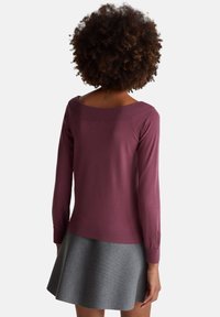 Žena s kudrnatými vlasy, oblečená v fialovém tričku s dlouhým rukávem a šedé sukni, stojící zády k divákovi na jednobarevném pozadí.