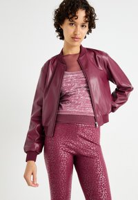 Veste bomber en cuir bordeaux avec des poignets côtelés, associée à un haut bordeaux ajusté avec un motif en script, et des leggings brillants à imprimé léopard.