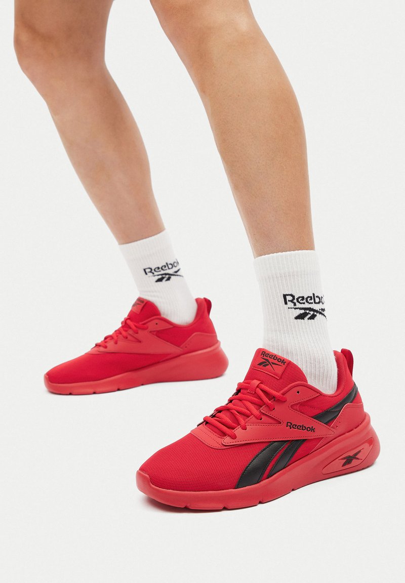 Červené športové topánky s mesh zvrškom, čiernymi akcentmi a hrubou podošvou. V kombinácii s bielymi ponožkami, ktoré zobrazujú logo Reebok.