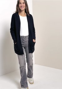 Marinblå stickad kofta med öppen front, två fickor, bärs över en vit skjorta och grå utsvängda jeans, parat med beige sneakers.