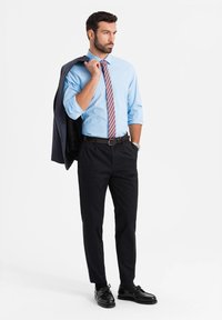 Abbigliamento da lavoro con una camicia azzurro chiaro, una cravatta a fantasia, pantaloni neri e scarpe nere lucide, completato da un blazer scuro appoggiato su una spalla.