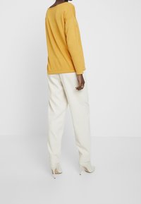 Hořčicově žlutý top s dlouhým rukávem a volným střihem, kombinovaný s bílými zúženými kalhotami a světle šedými lodičkami na vysokém podpatku. Jednoduchý, minimalistický design.