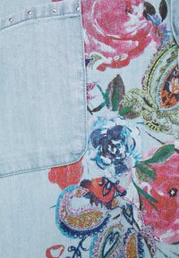 Tessuto in denim azzurro chiaro con motivi floreali e paisley colorati in rosa, rosso e verde, con dettagli metallici sulla tasca.