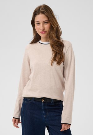 Jeune femme aux longs cheveux bruns portant un pull beige à manches longues, un jean bleu foncé et une ceinture noire, debout devant un fond uni.