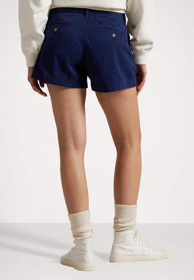 Marineblaue Baumwollshorts mit Gesäßtaschen und goldenen Knopfakzenten, kombiniert mit weißen High-Top-Sneakern und cremefarbenen gerippten Socken.