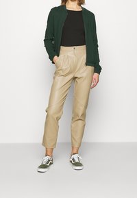 Veste bomber verte, haut côtelé noir, pantalon taille haute en similicuir beige, et baskets vert foncé et blanc, stylés avec des poches avant.