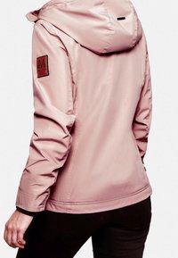 Femme portant une veste à capuche rose clair avec un écusson logo sur le haut de la manche gauche, vue de dos avec un pantalon noir.