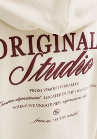 Kremasta kapuca z bordo napisom "ORIGINAL Studio" ter manjšimi frazami o ustvarjalnem oddelku in novih izrazih.