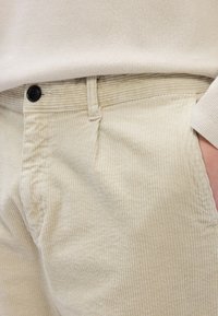 Calças de courino bege com riscas verticais, apresentando um fecho de botão preto e bolsos laterais, estilizadas com uma parte de cima de cor neutra.