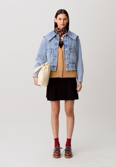 Veste en jean avec grand col, cardigan beige à boutons, robe noire plissée, chaussettes rouges, chaussures marron et sac à main crème.