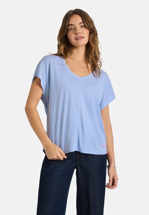 Woman with wavy hair wearing a light blue and white striped short-sleeve top and dark blue jeans, standing with one hand in pocket.