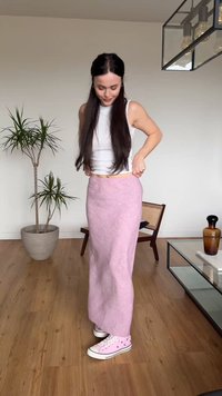 Kobieta poprawiająca długą różową spódnicę w pomieszczeniu, ubrana w biały bezrękawnik i różowe trampki, stoi na drewnianej podłodze obok rośliny i krzesła.