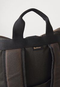 Close-up of black and brown backpack shoulder straps with a black handle and a small sewn-in label reading "Barbour."