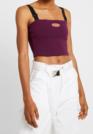 Top corto viola con spalline nere, presenta una toppa con logo al centro. Indossato con pantaloni bianchi con una cintura larga e tasche.