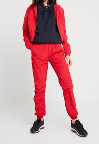 Röd bomberjacka med elastiska ärmar, ihopparad med matchande röda joggers med vita sidosträck. Svarta sportskor fulländar outfiten.