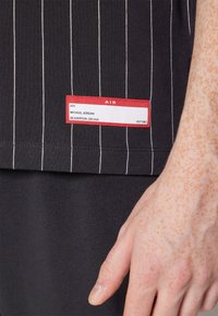 Primer plano del dobladillo de una camisa de rayas negras con una etiqueta roja y blanca que dice "#23 Michael Jordan Beaverton, OR USA" junto a una mano llena de pecas.