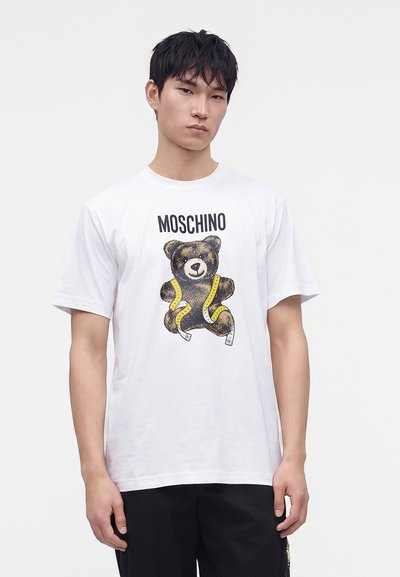 Νέος άνδρας που φορά ένα λευκό t-shirt Moschino με σχέδιο αρκουδάκι και κίτρινη μεζούρα, στέκεται μπροστά σε απλό φόντο.